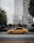 A yellow car is parked on the street in front of a modern building under construction, with a red crane visible in the background. There are trees along the side of the road and a sign for office space lease at the construction site.