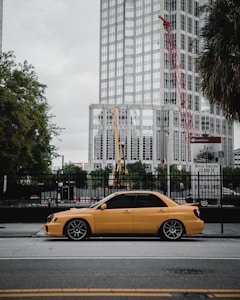 A yellow car is parked on the street in front of a modern building under construction, with a red crane visible in the background. There are trees along the side of the road and a sign for office space lease at the construction site.