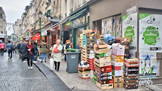A bustling city street with people walking along a cobblestone pathway. A stack of cardboard boxes and crates is placed near a building entrance, featuring signs for a coworking space. The street is lined with buildings, and some people appear to be engaged in conversation while others are simply passing by. Posters and advertisements can be seen on the walls.