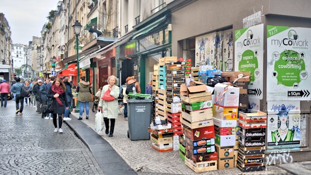 A bustling city street with people walking along a cobblestone pathway. A stack of cardboard boxes and crates is placed near a building entrance, featuring signs for a coworking space. The street is lined with buildings, and some people appear to be engaged in conversation while others are simply passing by. Posters and advertisements can be seen on the walls.