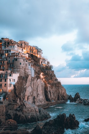 Several houses with pastel colors are perched on top of rugged cliffs overlooking the ocean. The setting sun casts a warm glow on the buildings, contrasting with the blue of the sea and sky. Waves crash against the rocks at the base of the cliff, and fluffy clouds are scattered across the sky.