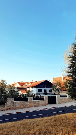 A suburban neighborhood features white buildings with red-tiled roofs surrounded by trees and stone fences. A wide road runs in the foreground, bordered by a red-and-white striped curb. The sky is clear and blue, suggesting a sunny day.