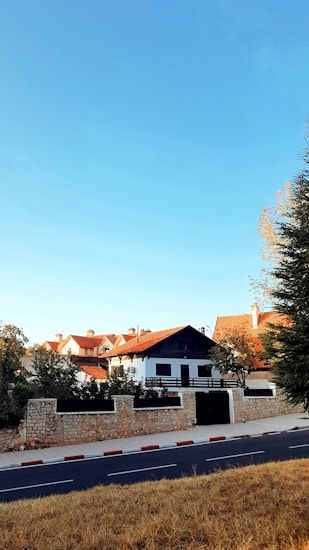 A suburban neighborhood features white buildings with red-tiled roofs surrounded by trees and stone fences. A wide road runs in the foreground, bordered by a red-and-white striped curb. The sky is clear and blue, suggesting a sunny day.