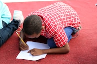 A child wearing a red and white checkered shirt leans over on a red carpeted floor, focused on writing with a pencil on a piece of white paper. Their legs are bent under them, and one shoe is visible to the left, belonging to another person sitting nearby.
