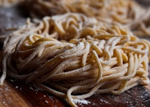 A close-up view of a pile of fresh, uncooked pasta noodles, sprinkled with flour. The image highlights the texture and details of the noodles, which appear to be homemade. The background is slightly blurred, emphasizing the focus on the pasta.