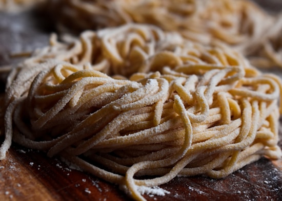 A close-up view of a pile of fresh, uncooked pasta noodles, sprinkled with flour. The image highlights the texture and details of the noodles, which appear to be homemade. The background is slightly blurred, emphasizing the focus on the pasta.