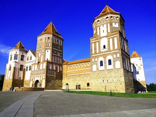 A large, historic castle with multiple towers made of brick and stone. The structure features decorative patterns on its walls and a pathway leading towards it. The architecture combines traditional elements with a striking symmetry. The sky is a vivid blue, providing a contrasting backdrop to the earthy tones of the castle.