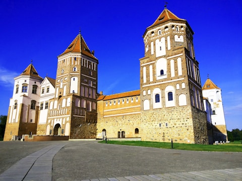 A large, historic castle with multiple towers made of brick and stone. The structure features decorative patterns on its walls and a pathway leading towards it. The architecture combines traditional elements with a striking symmetry. The sky is a vivid blue, providing a contrasting backdrop to the earthy tones of the castle.
