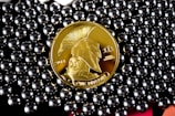 A gold-colored coin with a profile of a warrior wearing a helmet, labeled with '2014' and 'ONE BITCOIN', is surrounded by shiny, metallic beads.