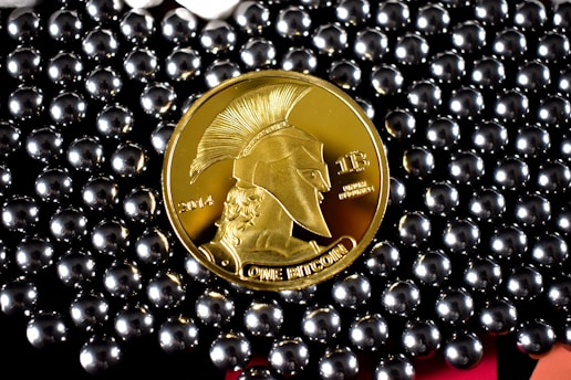 A gold-colored coin with a profile of a warrior wearing a helmet, labeled with '2014' and 'ONE BITCOIN', is surrounded by shiny, metallic beads.