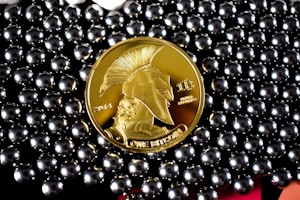 A gold-colored coin with a profile of a warrior wearing a helmet, labeled with '2014' and 'ONE BITCOIN', is surrounded by shiny, metallic beads.