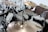 A drum set is positioned in a recording or rehearsal studio, featuring multiple cymbals, toms, and a bass drum. Microphones are set up around the drums, indicating a recording session. The room is modestly furnished with a chair visible in the background.