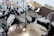 A drum set is positioned in a recording or rehearsal studio, featuring multiple cymbals, toms, and a bass drum. Microphones are set up around the drums, indicating a recording session. The room is modestly furnished with a chair visible in the background.