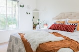 A brightly lit bedroom scene featuring a neatly made bed with a white and patterned bedspread, accented by a red and a white throw pillows. The bed has a beige tufted headboard, and a fluffy white throw is draped over it. On the wall, minimalistic decor includes round wall hangings with greenery. A modern white-framed mirror is positioned near a potted plant, adding a fresh element to the room. A window lets in natural light, enhancing the airy and serene atmosphere.