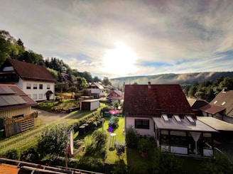 A peaceful rural neighborhood with several houses nestled amongst lush greenery. The sun is setting or rising, casting a warm light over the scene. There's a mix of traditional and modern architecture with well-maintained gardens.