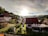 A peaceful rural neighborhood with several houses nestled amongst lush greenery. The sun is setting or rising, casting a warm light over the scene. There's a mix of traditional and modern architecture with well-maintained gardens.
