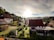 A peaceful rural neighborhood with several houses nestled amongst lush greenery. The sun is setting or rising, casting a warm light over the scene. There's a mix of traditional and modern architecture with well-maintained gardens.