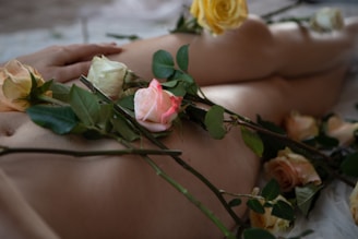 A nude form elegantly adorned with rose stems and lush green leaves, creating a soft and intimate atmosphere. The variety of roses includes shades of yellow, pink, and cream, enhancing the gentle and romantic feel. The play of light and shadow on the skin adds depth, while the positioning of the hand suggests a relaxed and peaceful state.