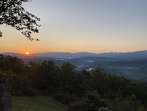 A serene landscape during sunrise with the sun appearing above a distant mountain range. The sky transitions from orange near the horizon to a clear blue further up. Mist drifts among the forested hills in the foreground, and a tree with extensive branches partially frames the view on the left side.