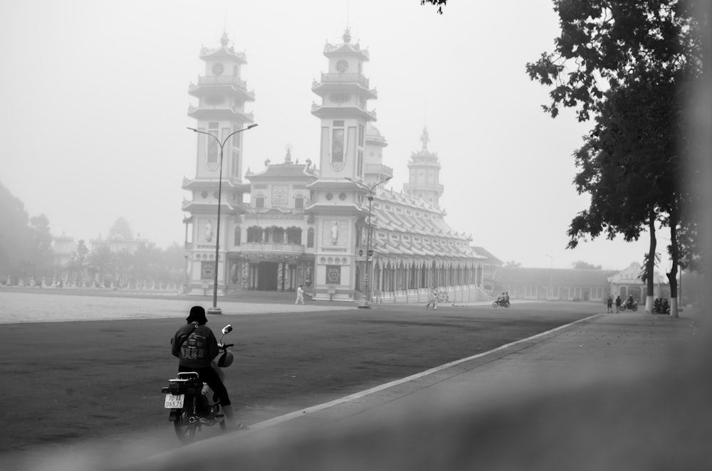 grayscale photo of man riding motorcycle on road near building