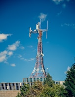 A telecommunications tower with a lattice structure stands tall against a clear blue sky. The tower is equipped with various antennas and dishes. Below the tower, the top of a modern building with a glass facade is visible, partially covered by green trees.