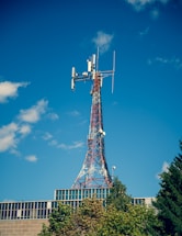 A telecommunications tower with a lattice structure stands tall against a clear blue sky. The tower is equipped with various antennas and dishes. Below the tower, the top of a modern building with a glass facade is visible, partially covered by green trees.
