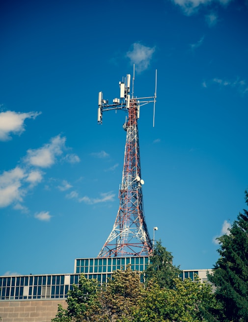 A telecommunications tower with a lattice structure stands tall against a clear blue sky. The tower is equipped with various antennas and dishes. Below the tower, the top of a modern building with a glass facade is visible, partially covered by green trees.