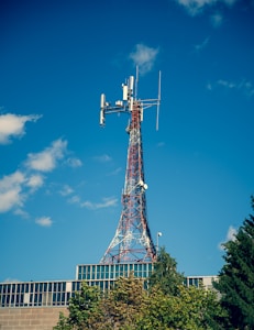 A telecommunications tower with a lattice structure stands tall against a clear blue sky. The tower is equipped with various antennas and dishes. Below the tower, the top of a modern building with a glass facade is visible, partially covered by green trees.