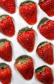 A collection of fresh, vibrant red strawberries neatly arranged in a white plastic tray, each nestled in its own compartment. The strawberries are plump with tiny seeds visible on their skin and fresh green leaves at the top. The surface of the strawberries appears glossy, indicating freshness, possibly with small dewdrops.