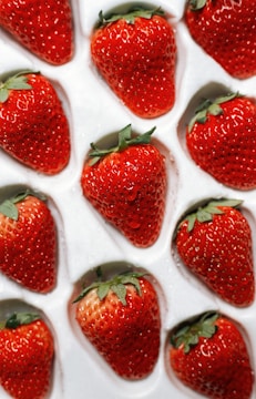A collection of fresh, vibrant red strawberries neatly arranged in a white plastic tray, each nestled in its own compartment. The strawberries are plump with tiny seeds visible on their skin and fresh green leaves at the top. The surface of the strawberries appears glossy, indicating freshness, possibly with small dewdrops.