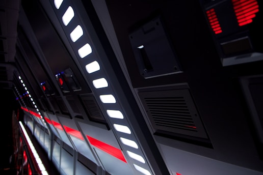 A futuristic and industrial corridor featuring angled metallic panels with bright white and red lights. The lights are embedded in a series of elongated rectangular vents and grooves, suggesting a high-tech environment.