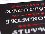 A close-up view of a printed page featuring the English alphabet in an ornate font style. The letters are displayed in white, while decorative elements surrounding them are in red. The background is black, creating a stark contrast that highlights the text and embellishments.
