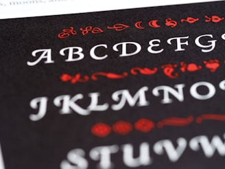 A close-up view of a printed page featuring the English alphabet in an ornate font style. The letters are displayed in white, while decorative elements surrounding them are in red. The background is black, creating a stark contrast that highlights the text and embellishments.