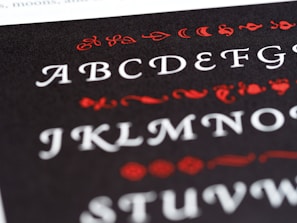A close-up view of a printed page featuring the English alphabet in an ornate font style. The letters are displayed in white, while decorative elements surrounding them are in red. The background is black, creating a stark contrast that highlights the text and embellishments.