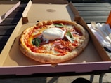 A freshly baked pizza with a crispy crust sits in an open cardboard box. The pizza is topped with melted cheese, slices of salami, red onions, and fresh basil leaves. A large dollop of burrata cheese is placed in the center. The box is placed on an outdoor wooden table beside a stack of disposable forks and napkins, with sunlight casting shadows across the scene.