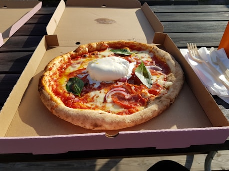 A freshly baked pizza with a crispy crust sits in an open cardboard box. The pizza is topped with melted cheese, slices of salami, red onions, and fresh basil leaves. A large dollop of burrata cheese is placed in the center. The box is placed on an outdoor wooden table beside a stack of disposable forks and napkins, with sunlight casting shadows across the scene.