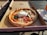 A freshly baked pizza with a crispy crust sits in an open cardboard box. The pizza is topped with melted cheese, slices of salami, red onions, and fresh basil leaves. A large dollop of burrata cheese is placed in the center. The box is placed on an outdoor wooden table beside a stack of disposable forks and napkins, with sunlight casting shadows across the scene.