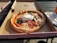 A freshly baked pizza with a crispy crust sits in an open cardboard box. The pizza is topped with melted cheese, slices of salami, red onions, and fresh basil leaves. A large dollop of burrata cheese is placed in the center. The box is placed on an outdoor wooden table beside a stack of disposable forks and napkins, with sunlight casting shadows across the scene.
