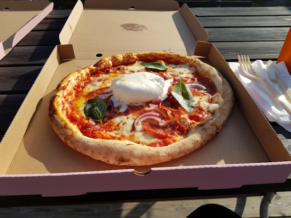A freshly baked pizza with a crispy crust sits in an open cardboard box. The pizza is topped with melted cheese, slices of salami, red onions, and fresh basil leaves. A large dollop of burrata cheese is placed in the center. The box is placed on an outdoor wooden table beside a stack of disposable forks and napkins, with sunlight casting shadows across the scene.