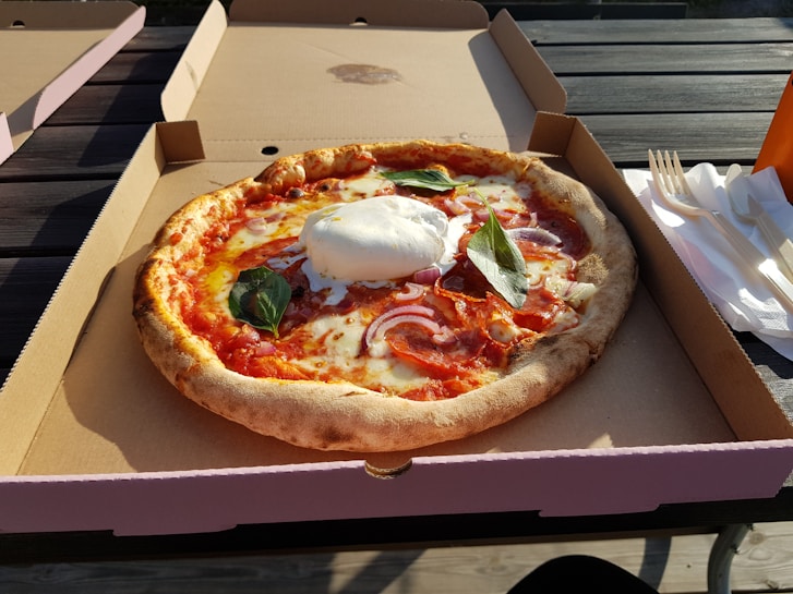 A freshly baked pizza with a crispy crust sits in an open cardboard box. The pizza is topped with melted cheese, slices of salami, red onions, and fresh basil leaves. A large dollop of burrata cheese is placed in the center. The box is placed on an outdoor wooden table beside a stack of disposable forks and napkins, with sunlight casting shadows across the scene.