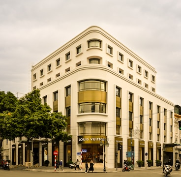A large, elegant corner building with multiple stories and sleek architectural design. The facade is adorned with the Louis Vuitton logo prominently displayed above the entrance. The building features large windows with gold-toned accents, creating a luxurious appearance. People and vehicles, including motorcycles, are visible on the street below.