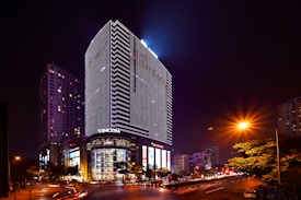 A night scene featuring a tall, modern skyscraper illuminated with bright, linear LED lights. The base of the building includes a shopping center with a visible 'VINCOM' sign. Surrounding the building are other structures, street lights, and trees, with light trails from passing vehicles contributing to a sense of motion.