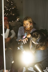 A group of people is gathered in a room, likely engaging in a tattoo session. A woman is holding a tattoo machine, working on someone’s arm or hand, while another person watches with interest. The setting is cozy with a decorated Christmas tree in the background, indicating a festive season.