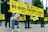 A group of people holding a large yellow banner with the words 'No More Factory Farms' are gathered in a public space. A dog in a blue and purple tie-dye sweater is standing in the foreground, adding a playful element to the scene. Other individuals in the background hold smaller, similar signs. The people appear to be dressed warmly, indicating possible cooler weather.
