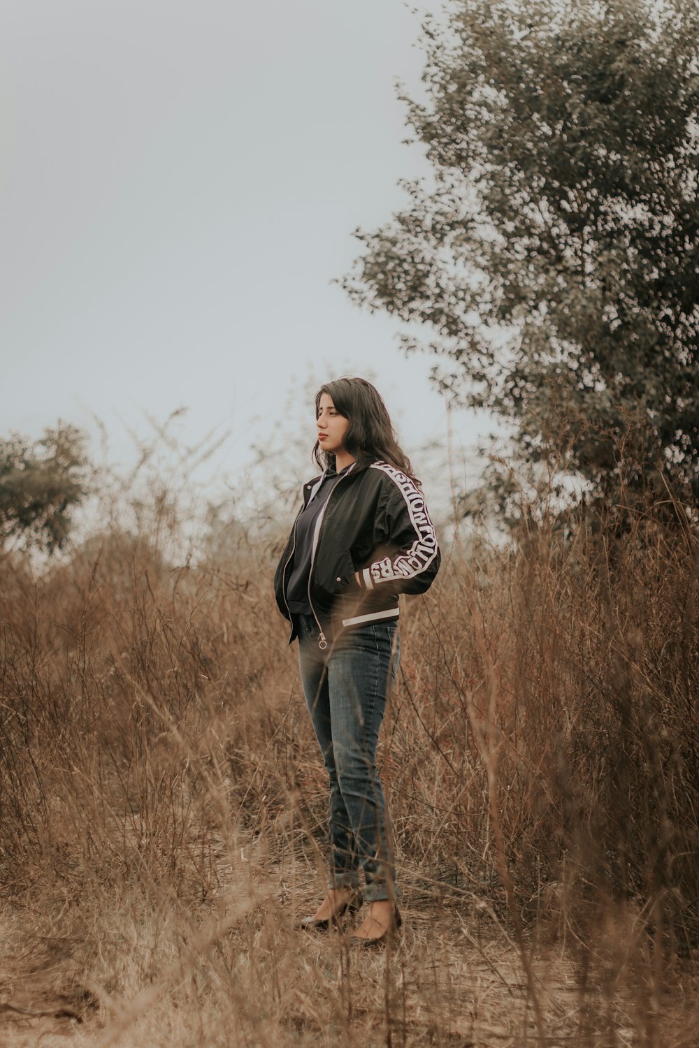 woman in black jacket and blue denim jeans standing on brown grass field during daytime