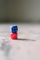Two dice are stacked one on top of the other on a smooth, reflective surface. The dice are colored differently, with the top one being blue and the bottom one red, each featuring white dots representing numbers. The background is out of focus, creating a soft and tranquil atmosphere.