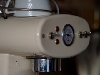 A close-up view of a vintage-style cream-colored espresso machine with a round temperature gauge indicating the temperature in degrees Celsius. The machine has two red indicator lights and silver toggle switches, and a metallic spout below. A white ceramic cup is partially visible on top.