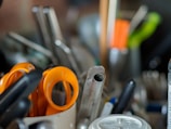 A collection of assorted utensils and tools, including visible orange-handled scissors, placed inside a container. The background is blurred, with other kitchen or office items present.