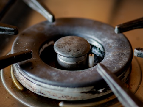 A close-up view of a gas stove burner with visible wear and tear, displaying rust and grime accumulated over time. The metal surface shows discoloration, and the central gas outlet is prominent.