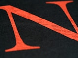 A large red letter N is overlaid on a black background, with small typed text visible within the letter. The text provides a textured appearance, and the color contrast is striking.
